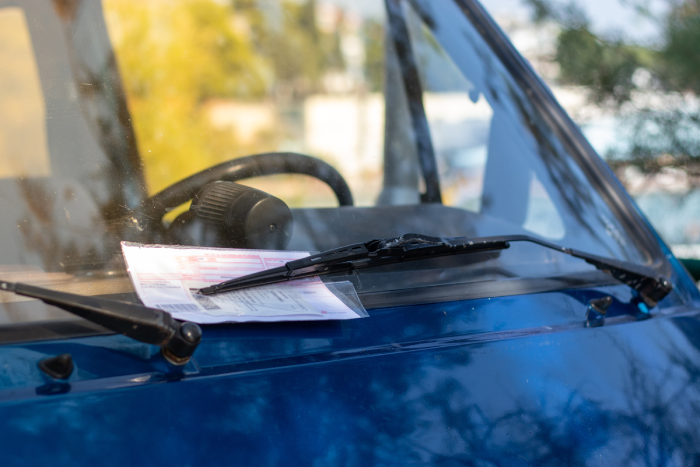Red parking ticket stuck under the windshielf wiper of a blue car, person fined for illegal parking in public.