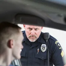 A male police officer with a stern expression, short light-colored hair, and a goatee is leaning slightly toward the window, making direct eye contact with the driver.