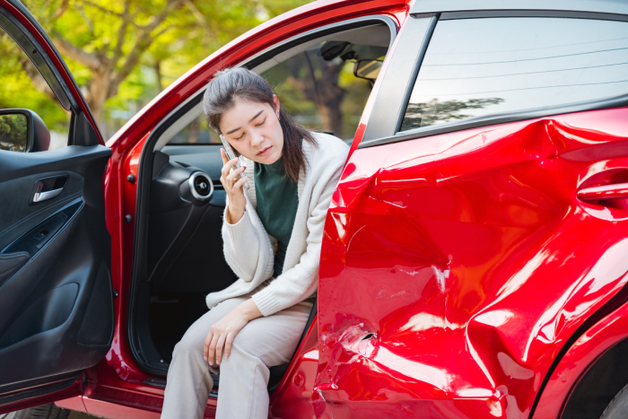 Car accident crash, Cars insurance protection, Claim collision, Auto emergency. A woman is sitting in a red car with a smashed front bumper. She is talking on her cell phone.