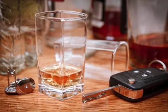 Gemini said A shot glass containing a small amount of amber-colored alcohol sits on a wooden surface next to a car key with the blade extended, symbolizing the dangers of drinking and driving. Multiple bottles and another glass are visible in the blurred background.