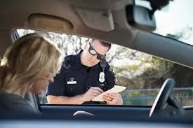 The officer is leaning toward the window while writing on a ticket or notepad. The back of the driver's head, a woman with blonde hair, is visible in the foreground.