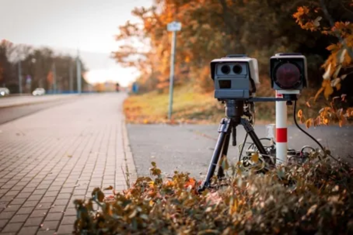 The image shows a mobile speed camera system set up on a tripod next to a road during autumn.