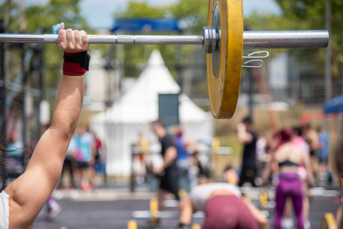 Scene of an outdoor weightlifting competition, with an athlete lifting a barbell with yellow discs in the foreground,